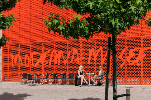 Besökare i caféstolar framför Moderna Museet Malmös orangea fasad.