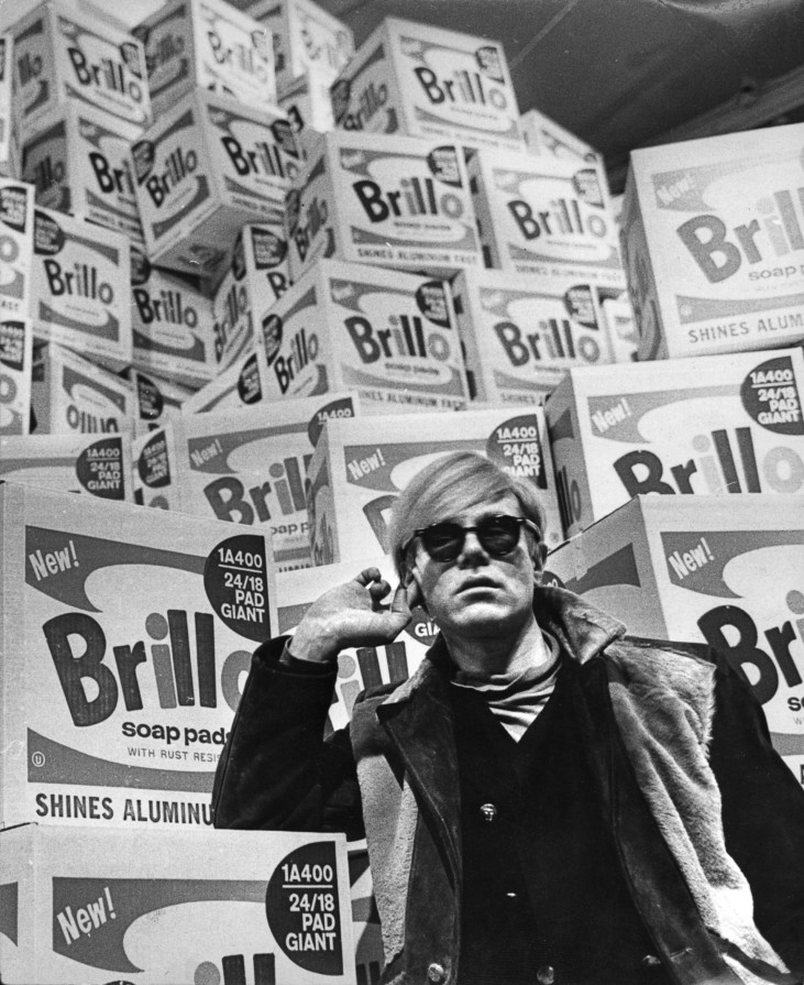 Andy Warhol standing in front of pile of Brillo boxes.