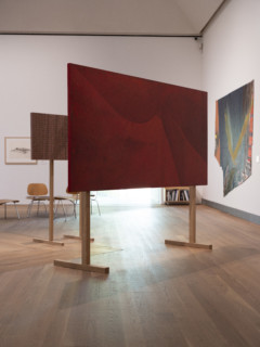 Image of a red canvas placed on wooden stands. In he background: chairs, artworks hung on white walls. 