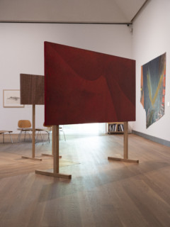 Image of a red canvas placed on wooden stands. In he background: chairs, artworks hung on white walls.