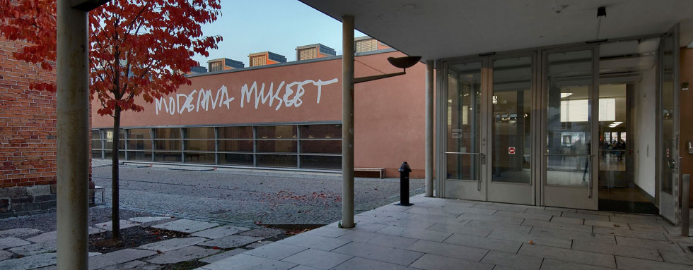Roof held up by pillars in front of two double doors. Tree with red leaves visible to the left of the entrance.