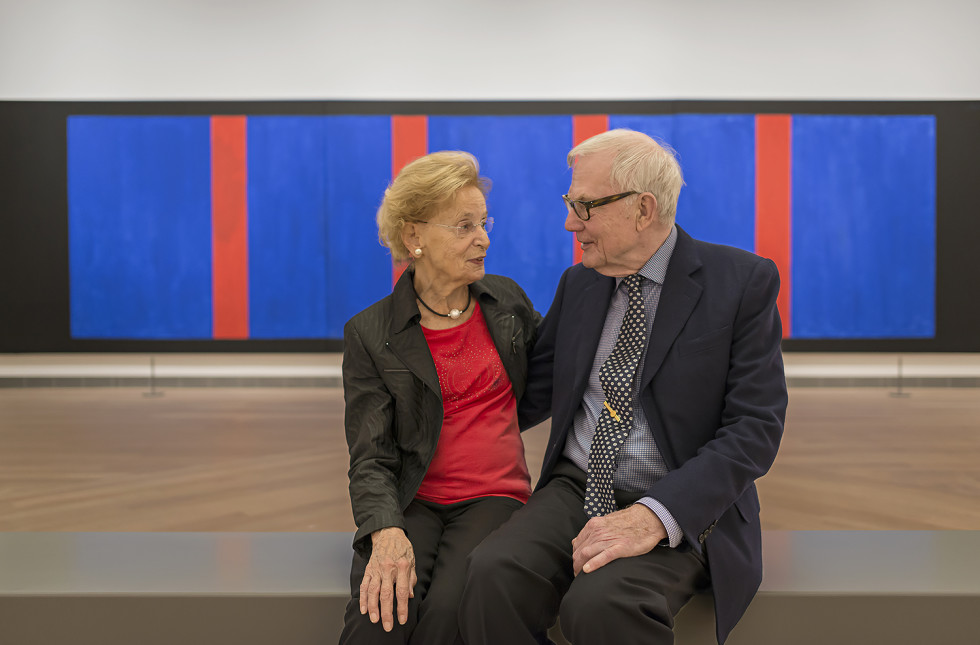 Two elderly people sitting in front of art work in the collection.