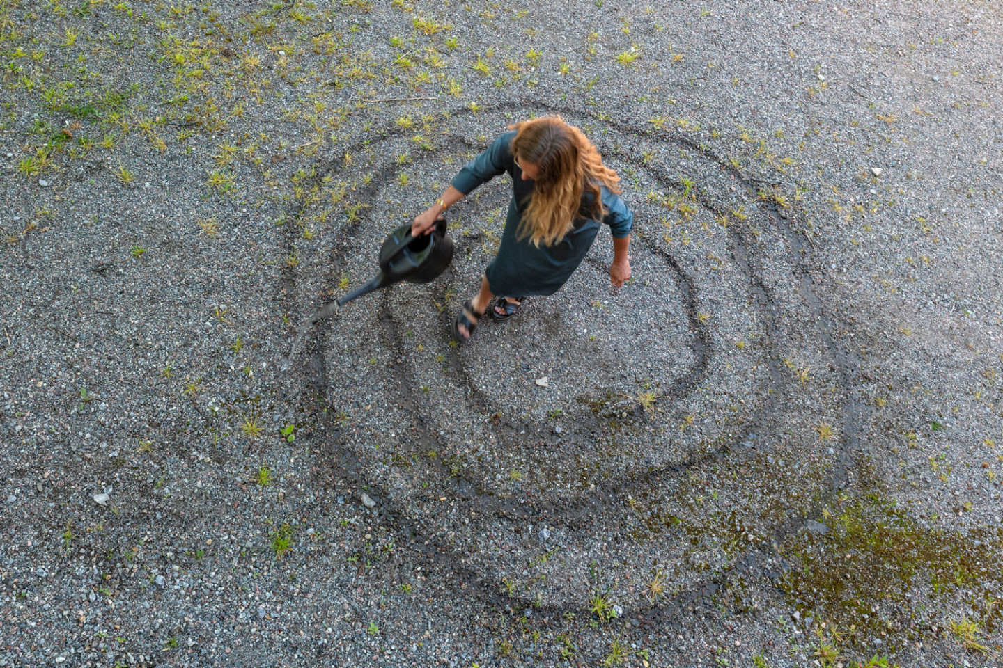 A person with long hair pours water around themself in a spiral with a watering can.