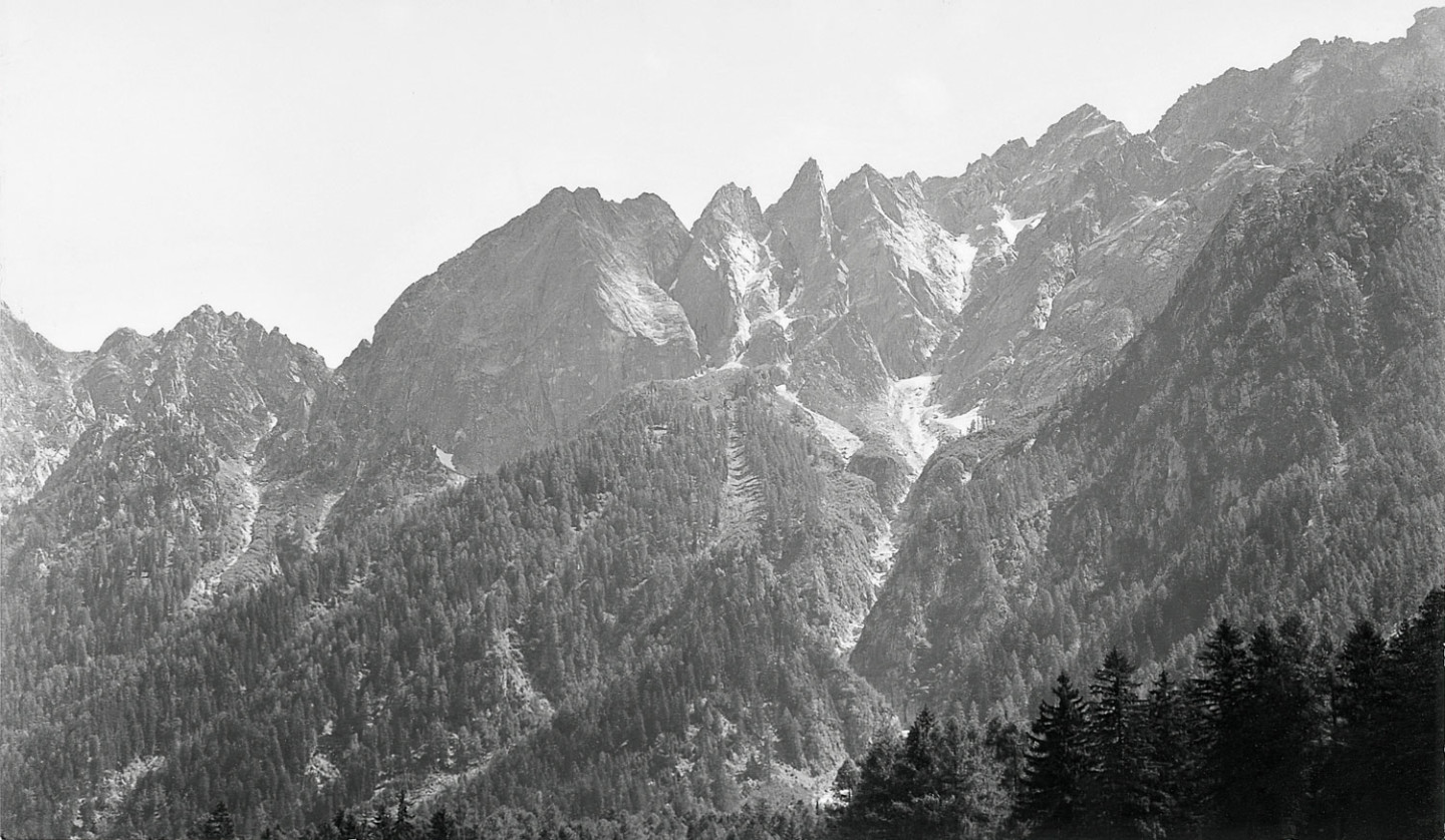Svartvitt fotografi av branta bergstoppar med skog längst ner.