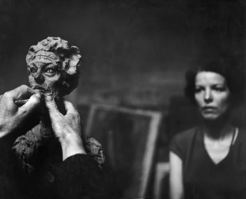 Hands sculpting in clay, woman sitting in the background, out of focus.