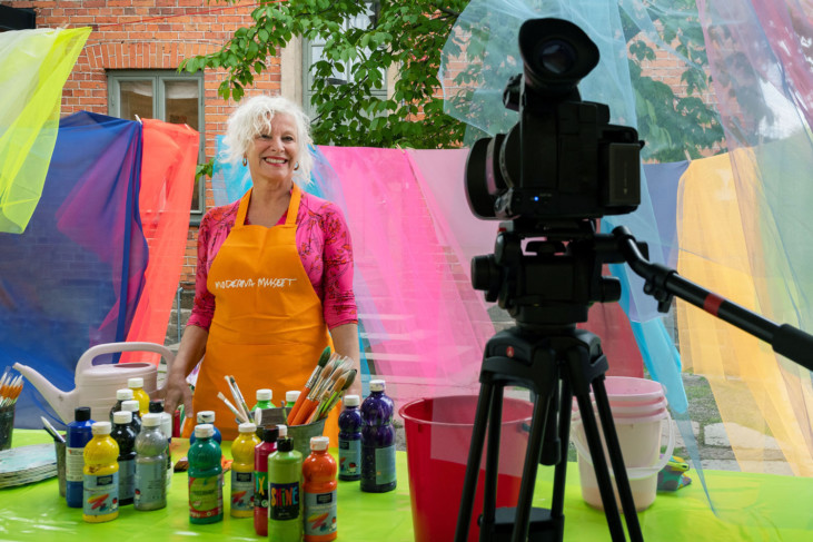 Smiling person standing in front of table with color tubes. Film camera visible in the foreground.