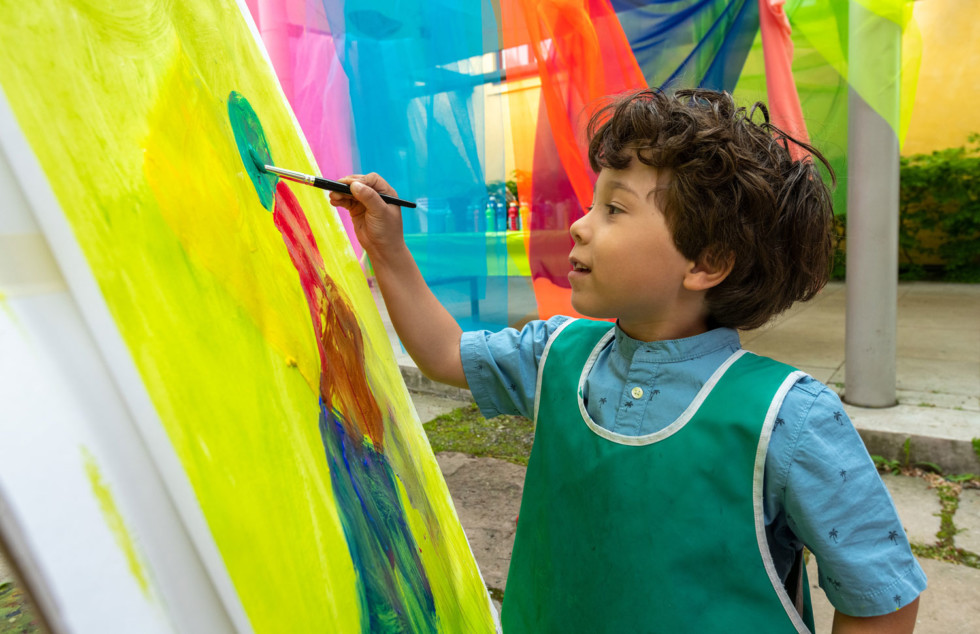 Children with brush in hand painting on canvas on easel.