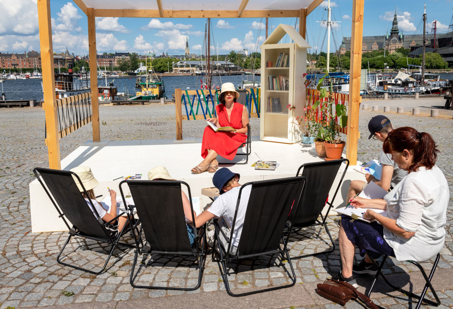 A person in a red dress sits on a small stage and reads to children and adults who paint.