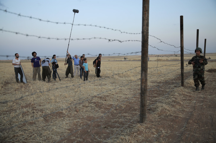 A group of people are filming out in a field and a barbed wire fence can be seen in the foreground. On the other side of the barbed wire is a soldier.