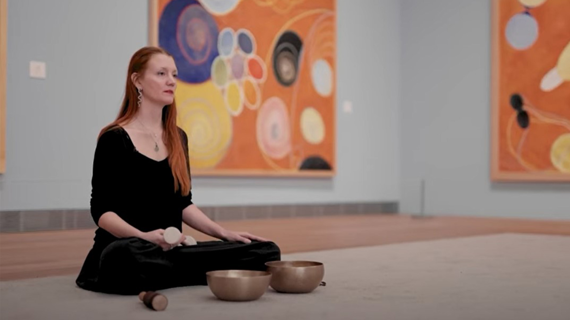Meditation guide Jessica Eldenstjärna is sitting with crossed legs on the floor in front of some of Hilma af Klint's paintings from the series "The Ten Largests"