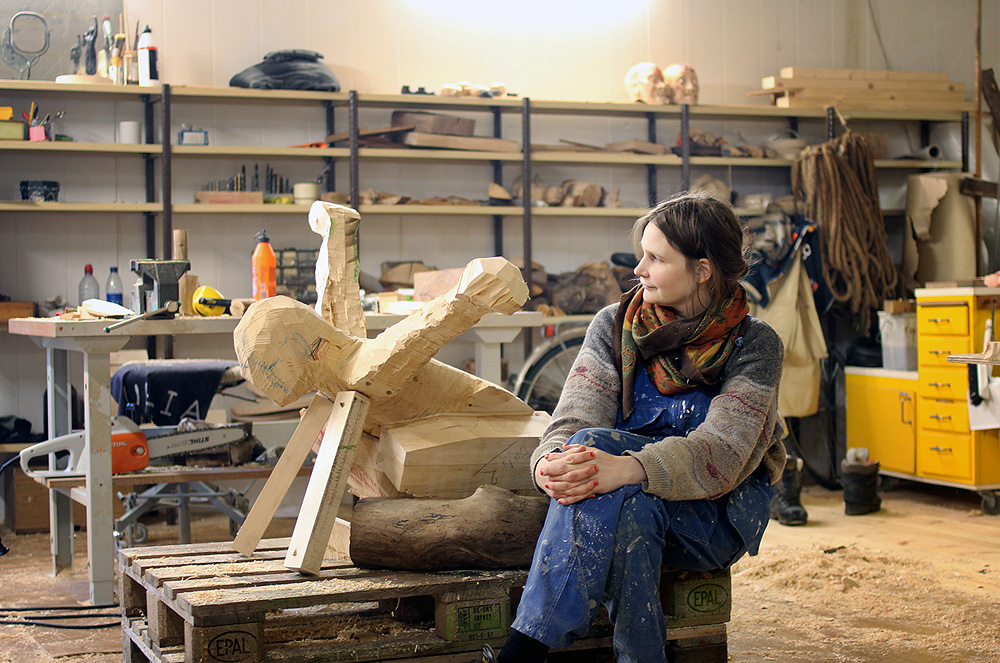 The artist sits and looks at a wooden sculpture.