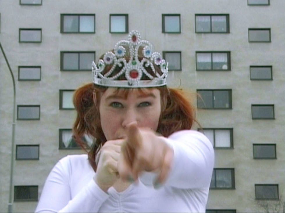 A person with a princess crown on his head points straight into the camera