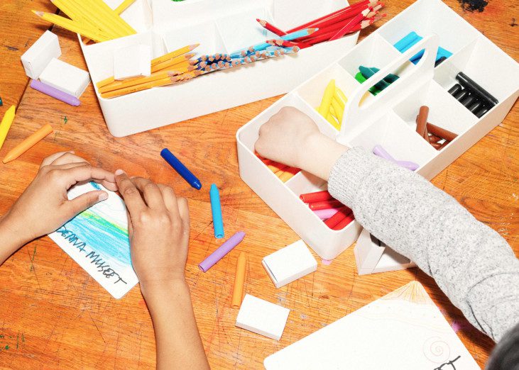 Photo of table with crayons and paper, two childrens' hands are reaching for crayons and coloring
