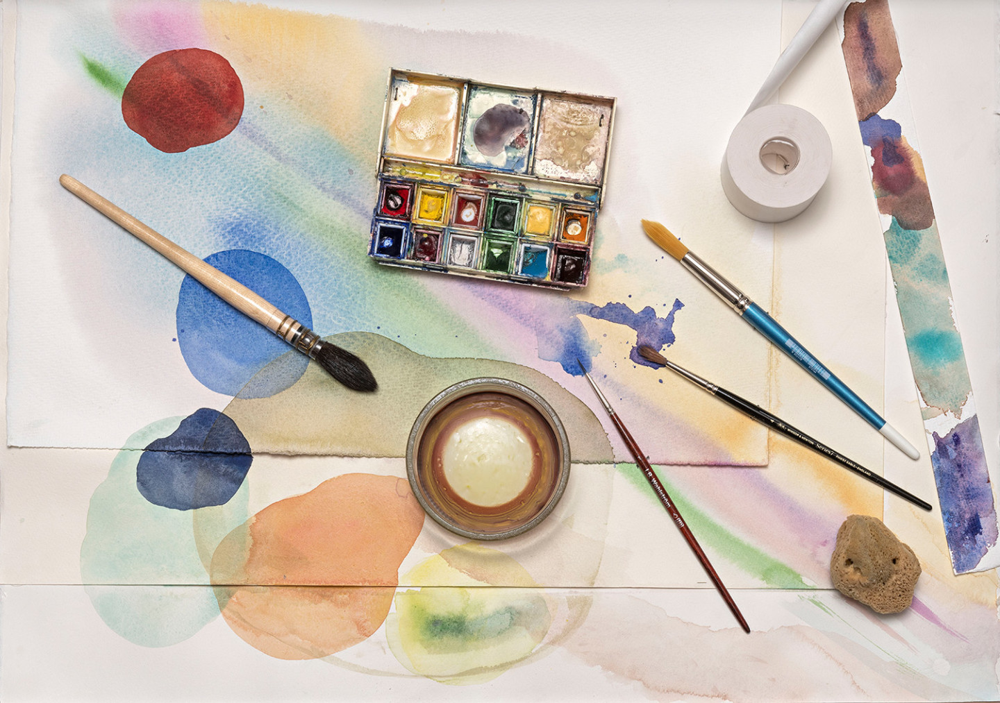 Watercolor painting with circles in different colors. There are brushes, a palette, masking tape and a glass of water placed around the painting.