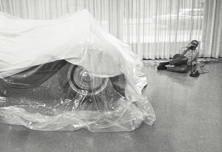 Pontus Hultén is lying on the floor and talking on the phone. In the foreground is a car covered with plastic