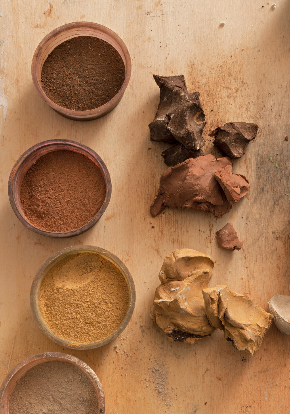 Three round containers with color pigments made from different clays, in the colors brown, red and beige. Next to the containers there are three lumps of clay, in the same colors.