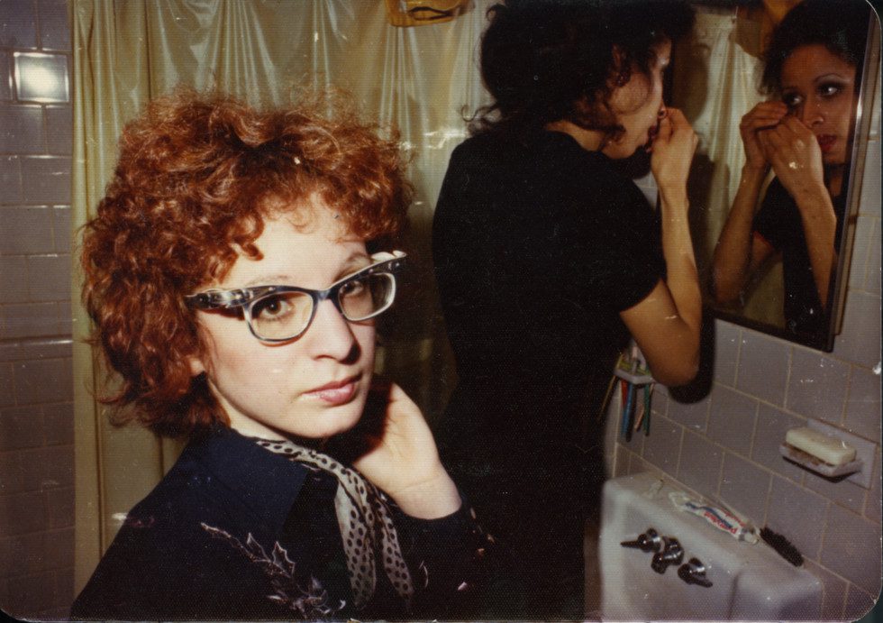 Nan Goldin stands in a bathroom with another person looking at herself in the mirror