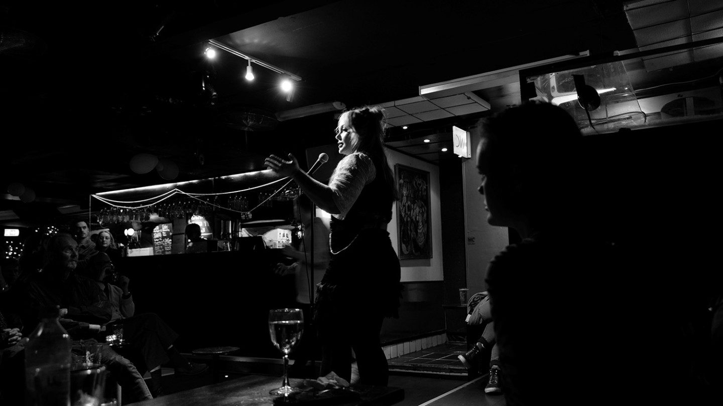 Black and white photograph of a person standing on a stage, talking into a microphone