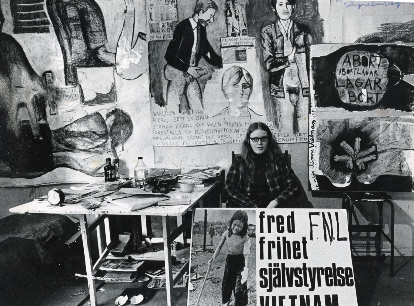Svart vit fotografi med Monica Sjöö omgiven av målningar och plakat
