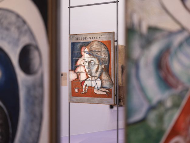 Installation view over the exhibition. Artworks are displayed on metal pipes. Behind two artworks we see the painting "House–Wives". The painting depicts a big head, a figure omn it's knees scrubbing the floor and a naked female body. Colorblocks in red, blue and beige. Over all figures is a grid.