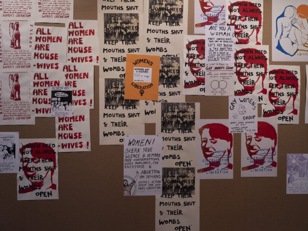 Photograph of prints in A4-format with repeated graphic prints nailed to a bulletin board. The prints read "all women are house-wives", "womens liberation" and "women need not always keep their mouth shut and their wombs open".