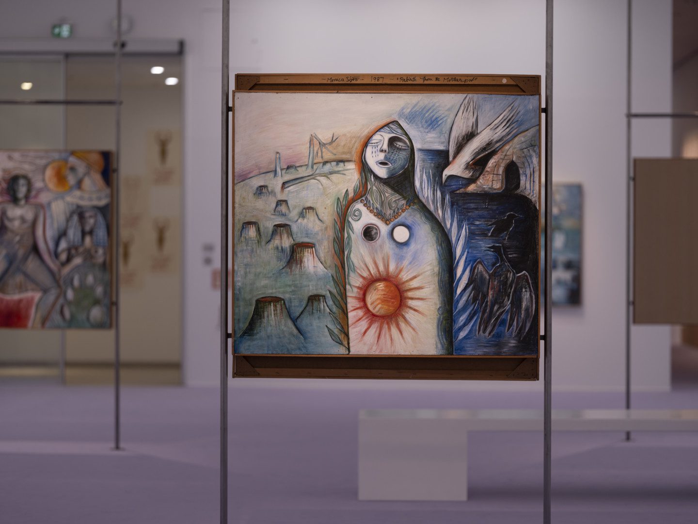 Photography of part of the exhibition area. In a purple room, the painting "Mother Earth in Pain, Her Trees Cut Down" is placed on narrow iron pipes. In the panting, a figure with a sun on the stomach is surrounded by chopped down trees in green on one side, and on the other a dark landscape with two black birds, one white bird and a cave.