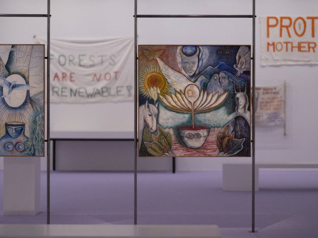 Photography of part of the exhibition area. In a purple room, the painting "Rebirth from the Motherpot" is placed on narrow iron pipes. In the painting, various figures are gathered around a pot with a blossomed yellow flower in it. The painting is in yellow, blue, red and dark blue.