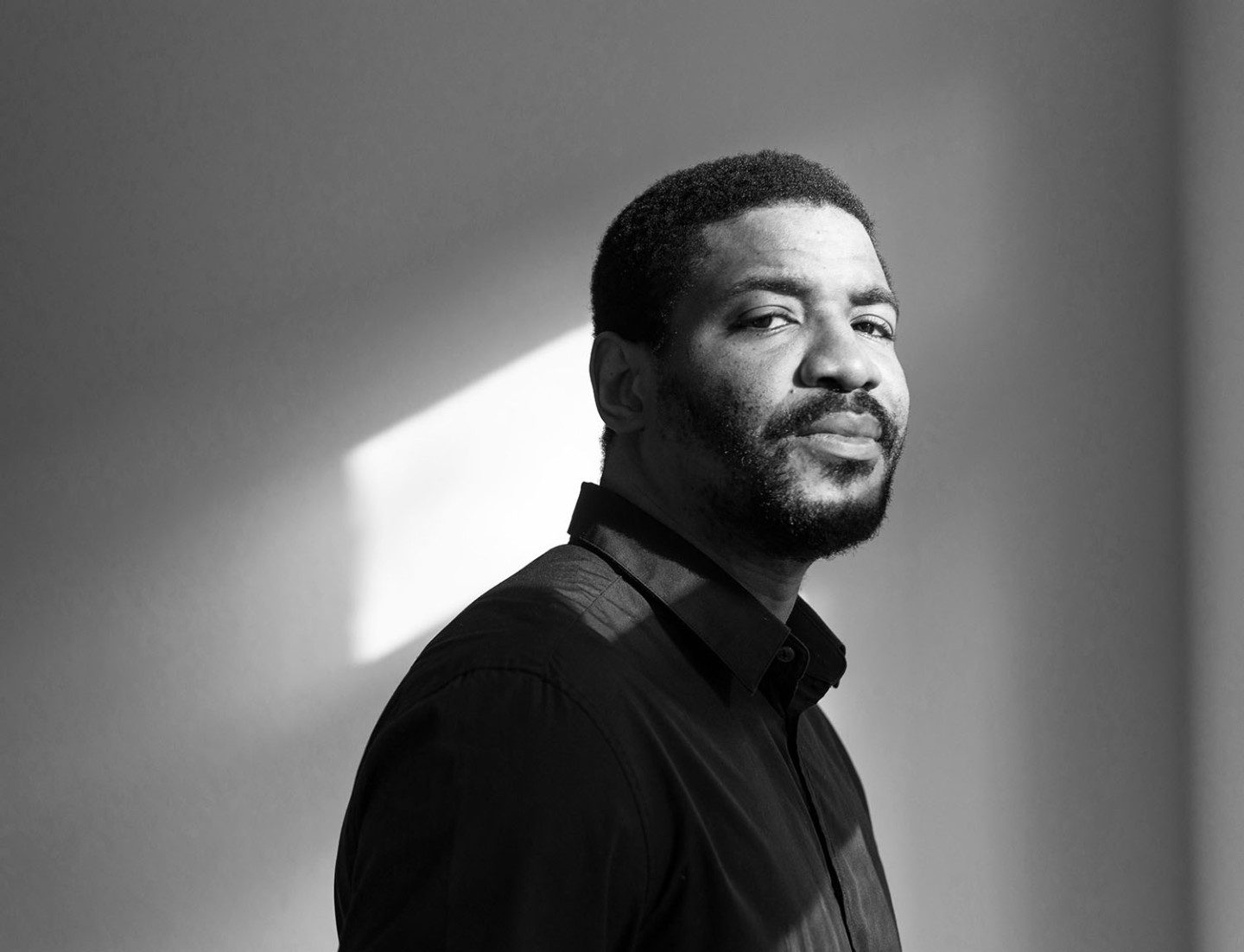 Black and white photo of the artist Emeka Ogboh, dressed in a dark shirt, looking at the camera from the right