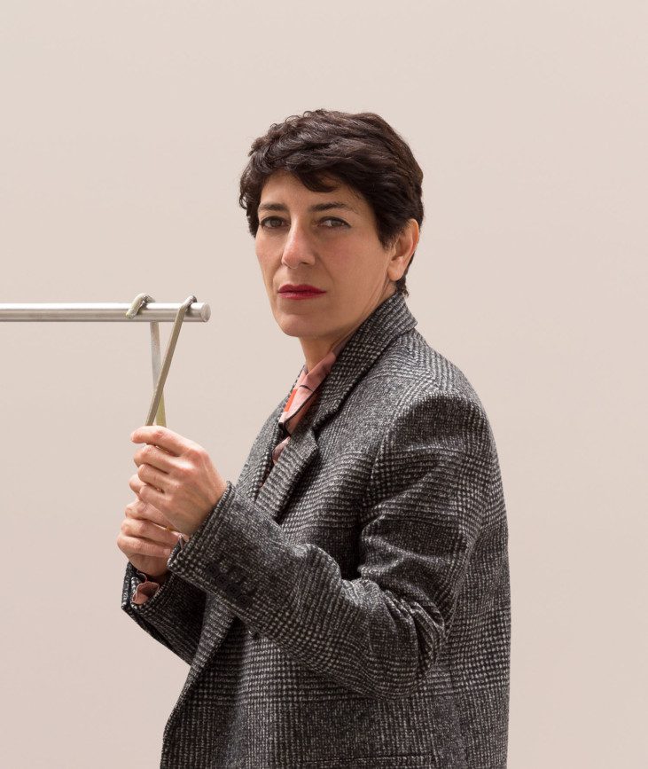 Photo of artist Nairy Baghramian, looking towards the camera in a grey blazer, white background