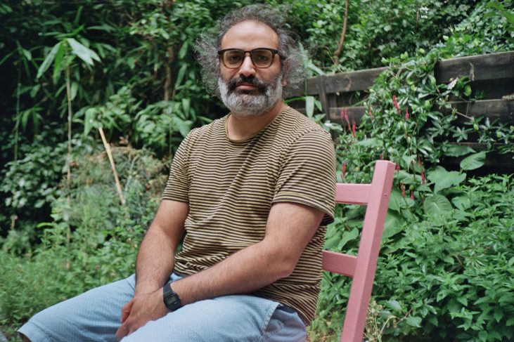 Photo of Yazan Khalili, sitting on a wooden chair with vegetation in the background