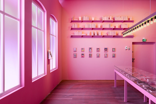 Installation view from the room "The Wicked Pavilion". A room completely in pink, with a stand and shelves with books on the wall in the picture. Paintings hang under the shelves, and three church-style windows can be seen next to the wall.
