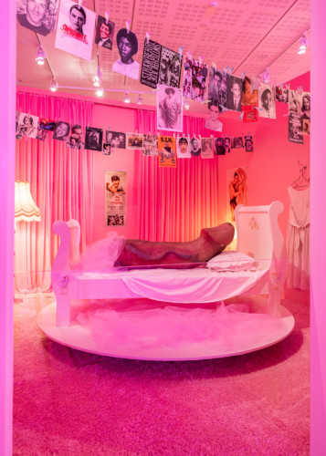 Installation view from the room "The Wicked Pavilion". Photograph of a girl's room in pink. A fishing line with photographs hangs from the ceiling. In the middle of the room is a bed on a podium, in the bed is a large phallus sculpture. 