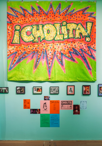 Installation view from the room "The Carla DuPlantier Cinerama Dome". Photograph on a wall of photographs and prints of or about Vaginal Davis. Above hangs a cloth with the words "CHOLITA" on it.