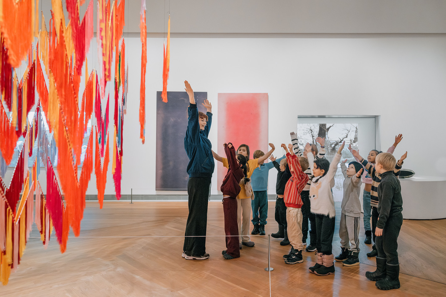 Barn sträcker upp armarna tillsammans med konstpedagog intill färgglada konstverk i en utställningssal